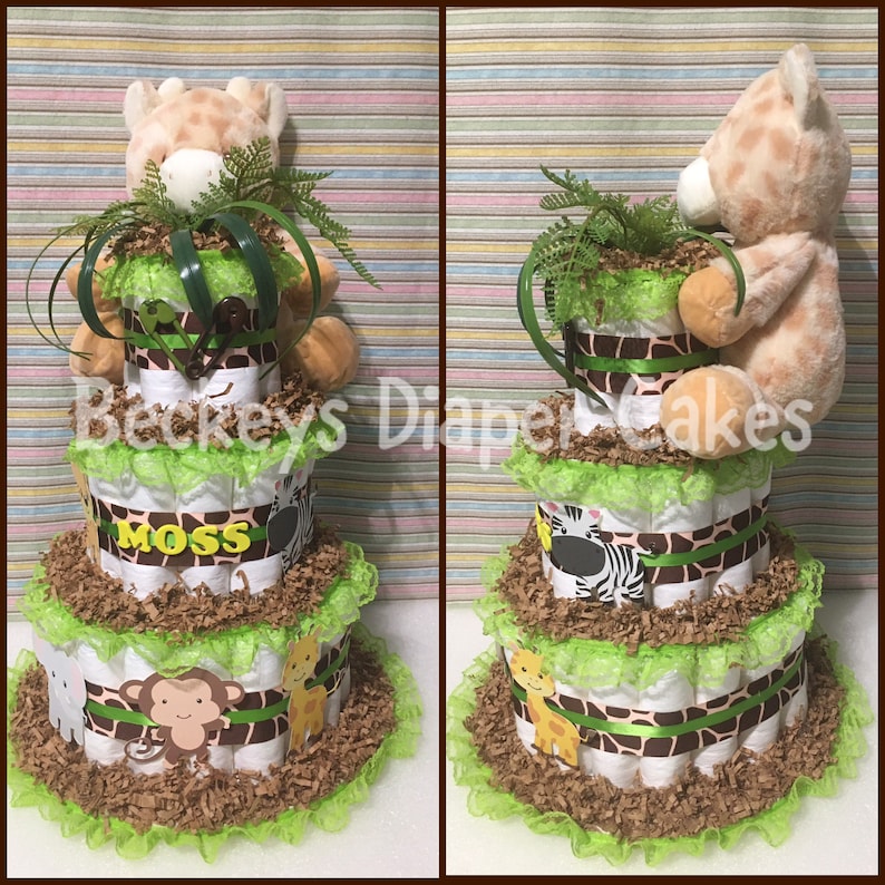 Op de afbeelding: Een drielaagse luiercake versierd met een giraffen-, zebra- en apenprint. De cake is versierd met groene en bruine papieren strooisel en groene kant. De bovenste laag heeft een knuffelgiraf erop. De middelste laag heeft een bruin lint met de naam "Moss" erop.
