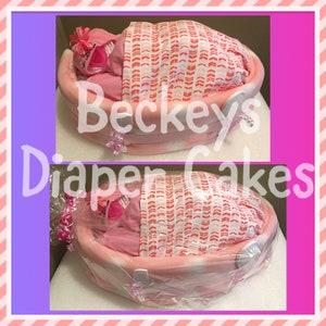 May include: Pink diaper cake shaped like a baby crib with a pink blanket and a pink baby doll inside. The cake is decorated with pink and white bows and the text "Beckeys Diaper Cakes".