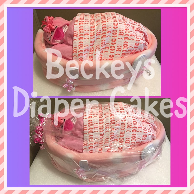 May include: Pink diaper cake shaped like a baby crib with a pink blanket and a pink baby doll inside. The cake is decorated with pink and white bows and the text "Beckeys Diaper Cakes".