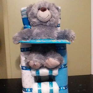 Op de afbeelding: Een grijze teddybeer zit op een blauwe en witte luiercake. De cake is versierd met blauwe en witte linten en de woorden "Baby" in blauw.