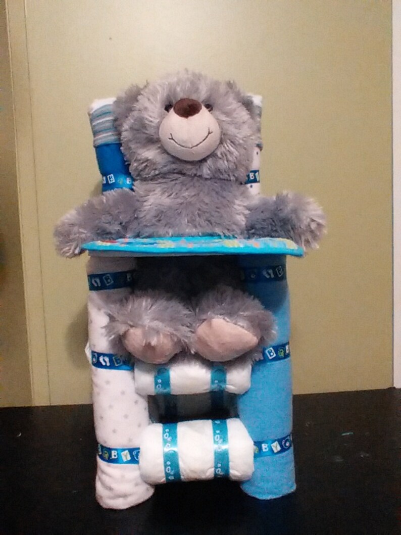 Op de afbeelding: Een grijze teddybeer zit op een blauwe en witte luiercake. De cake is versierd met blauwe en witte linten en de woorden "Baby" in blauw.