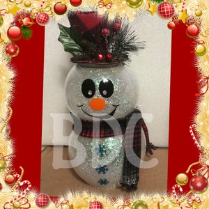 Peut inclure: Un bonhomme de neige blanc paillet&eacute; avec un chapeau et une &eacute;charpe en tartan rouge et noir. Le bonhomme de neige a des yeux noirs, un nez orange et une bouche souriante.