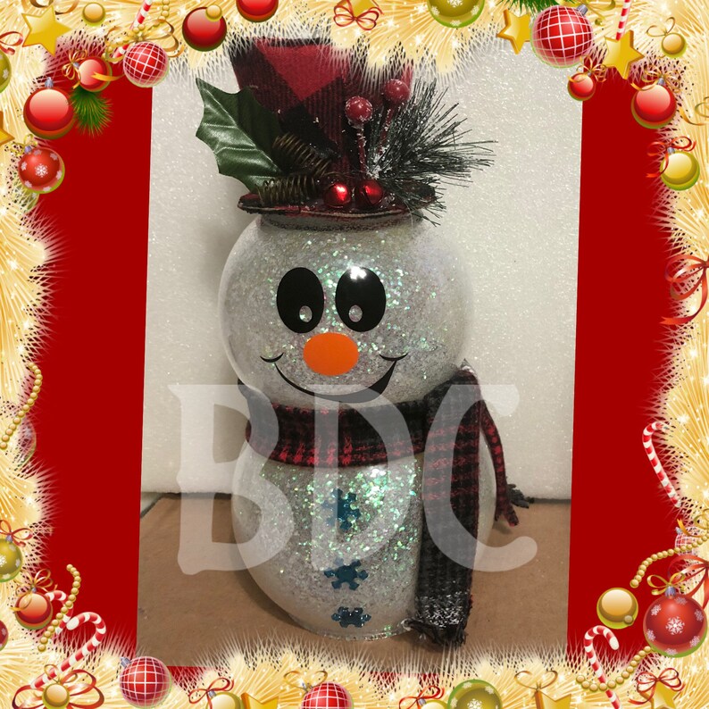 Peut inclure: Un bonhomme de neige blanc paillet&eacute; avec un chapeau et une &eacute;charpe en tartan rouge et noir. Le bonhomme de neige a des yeux noirs, un nez orange et une bouche souriante.
