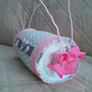May include: A grey and white nappy cake with pink ribbon and bows. The cake is wrapped in a grey and white chevron pattern fabric and has a pink polka dot ribbon around it. The cake is decorated with pink bows and is held together with a pink ribbon.