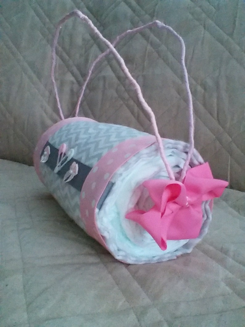 May include: A grey and white nappy cake with pink ribbon and bows. The cake is wrapped in a grey and white chevron pattern fabric and has a pink polka dot ribbon around it. The cake is decorated with pink bows and is held together with a pink ribbon.