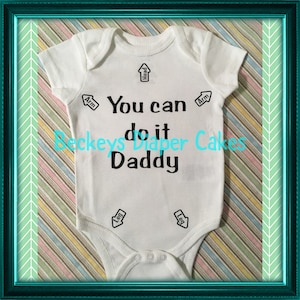 May include: White baby onesie with black text that reads "You can do it Daddy" with arrows pointing to "Head", "Arm", "Leg" and "Leg".