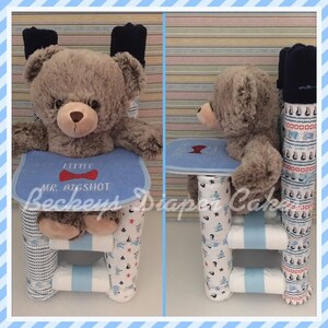Op de afbeelding: Een blauwe en witte luiercake in de vorm van een hoge stoel met een teddybeer die erin zit. De cake is versierd met een blauw-wit nautisch thema en de teddybeer draagt ​​een blauwe vlinderdas. De cake is gelabeld "Little Mr. Bigshot".