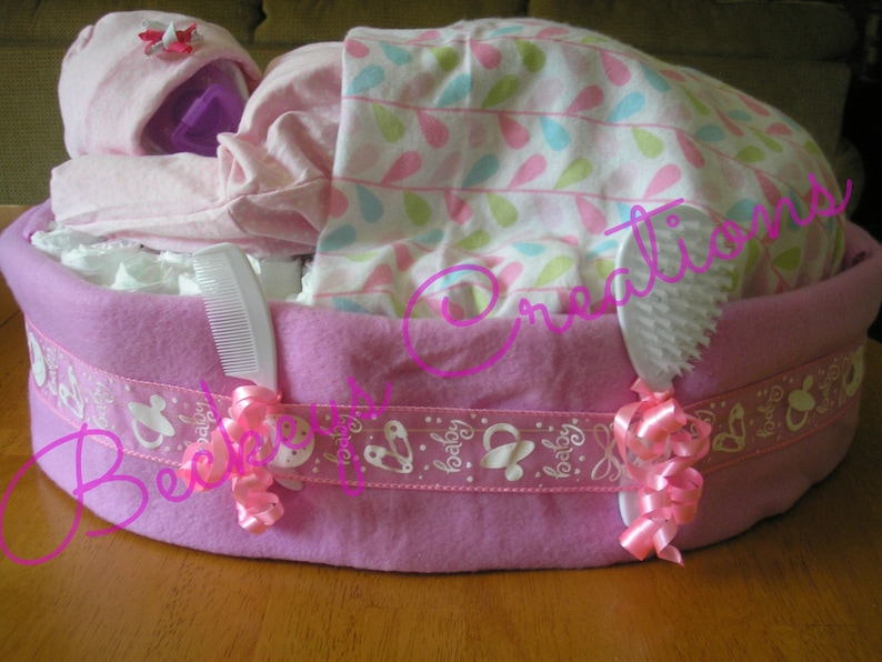 May include: A pink and white diaper cake with a pink blanket, a pink hat, and a white comb. The cake is decorated with a pink ribbon and the words "Baby" in white.