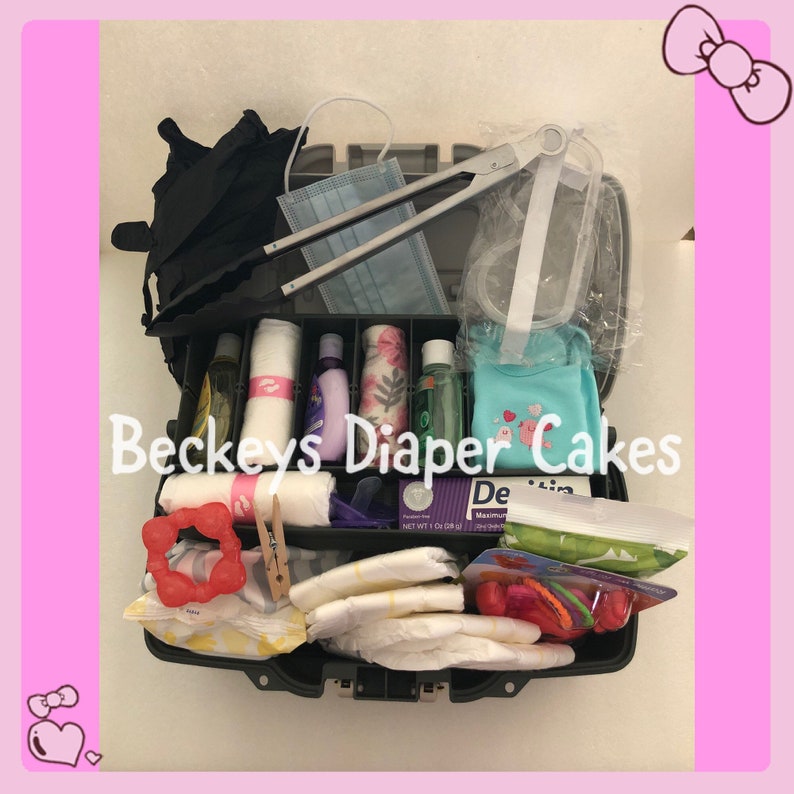 May include: A black plastic toolbox filled with baby items for a diaper cake. The items include nappies, wipes, lotion, a baby outfit, a dummy, a teether, and a pair of tongs. The toolbox has the text "Beckeys Diaper Cakes" printed on the side.