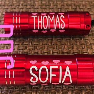 May include: Two red metal flashlights with white lettering and pink hearts. The top flashlight says "THOMAS" and the bottom flashlight says "SOFIA".