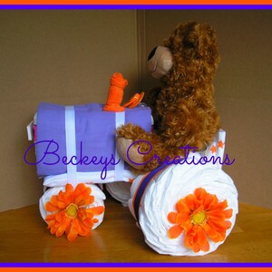 May include: A purple and white diaper cake shaped like a tractor with a brown teddy bear sitting on top. The tractor has orange wheels and flowers. The cake is decorated with the text "Beckeys Creations".