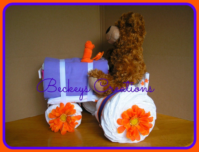 May include: A purple and white diaper cake shaped like a tractor with a brown teddy bear sitting on top. The tractor has orange wheels and flowers. The cake is decorated with the text "Beckeys Creations".
