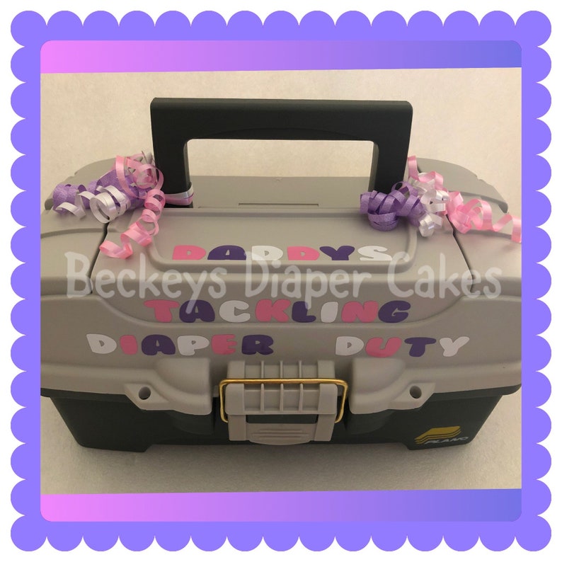 May include: A grey plastic toolbox with a black handle and gold latch. The toolbox has pink, purple and white letters that say "Daddy's Tackling Diaper Duty" and "Beckeys Diaper Cakes".