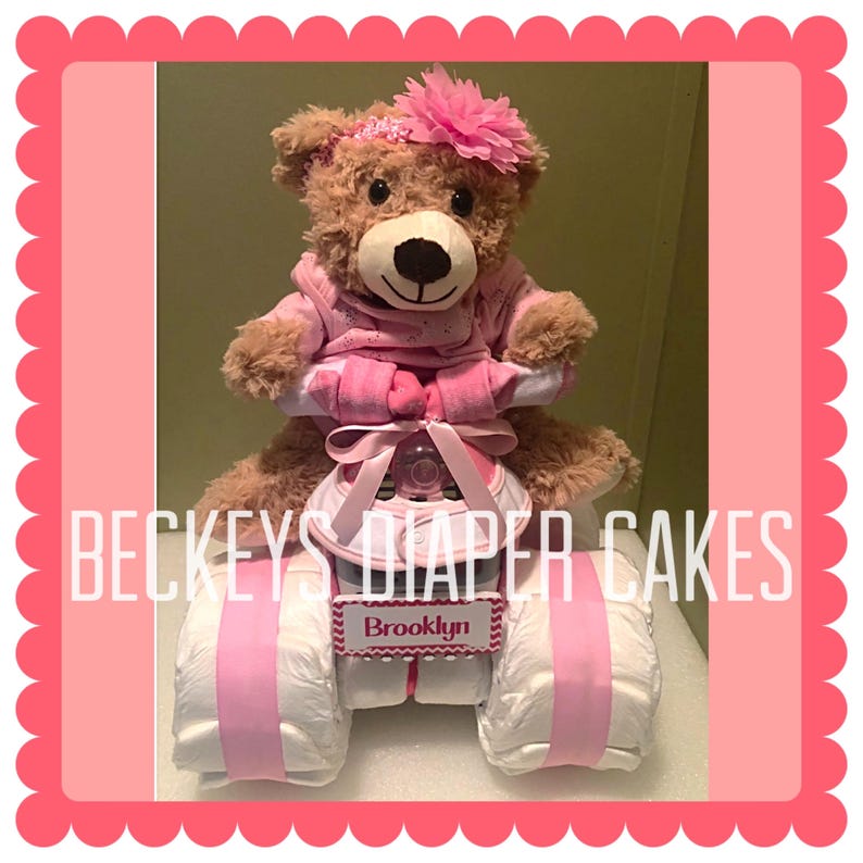 Pu&ograve; includere: Una torta di pannolini rosa e bianca a forma di quad a quattro ruote con un orsacchiotto di peluche che indossa un vestito rosa seduto sul sedile. La torta &egrave; decorata con un nastro rosa e un cartello che dice "Brooklyn".