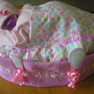 May include: A pink diaper cake with a baby doll, a white baby brush, and pink ribbons. The cake is decorated with a white and pink patterned fabric and a pink ribbon with the word "Baby" on it.