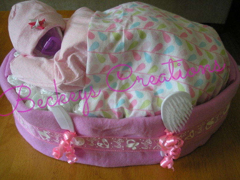 May include: A pink diaper cake with a baby doll, a white baby brush, and pink ribbons. The cake is decorated with a white and pink patterned fabric and a pink ribbon with the word "Baby" on it.