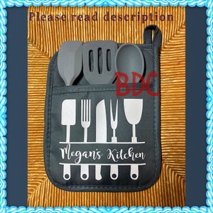 May include: A black kitchen utensil holder with a white design of kitchen tools and the text "Megan's Kitchen". The holder has a pocket on the front to hold utensils. The holder is hanging on a brown wicker surface.