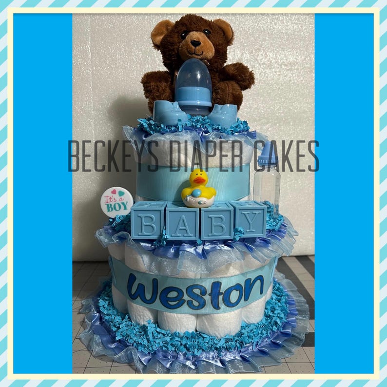 May include: A three-tiered diaper cake in shades of blue, decorated with a brown teddy bear, baby bottle, and baby shoes. The cake features the text "BECKEYS DIAPER CAKES", "It's a Boy" and "Weston". The top tier has blocks that spell "BABY".