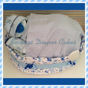 May include: A blue and white diaper cake decorated with a dog theme. The cake is in a basket with a blue blanket on top. The text "Beckeys Diaper Cakes" is visible on the blanket.