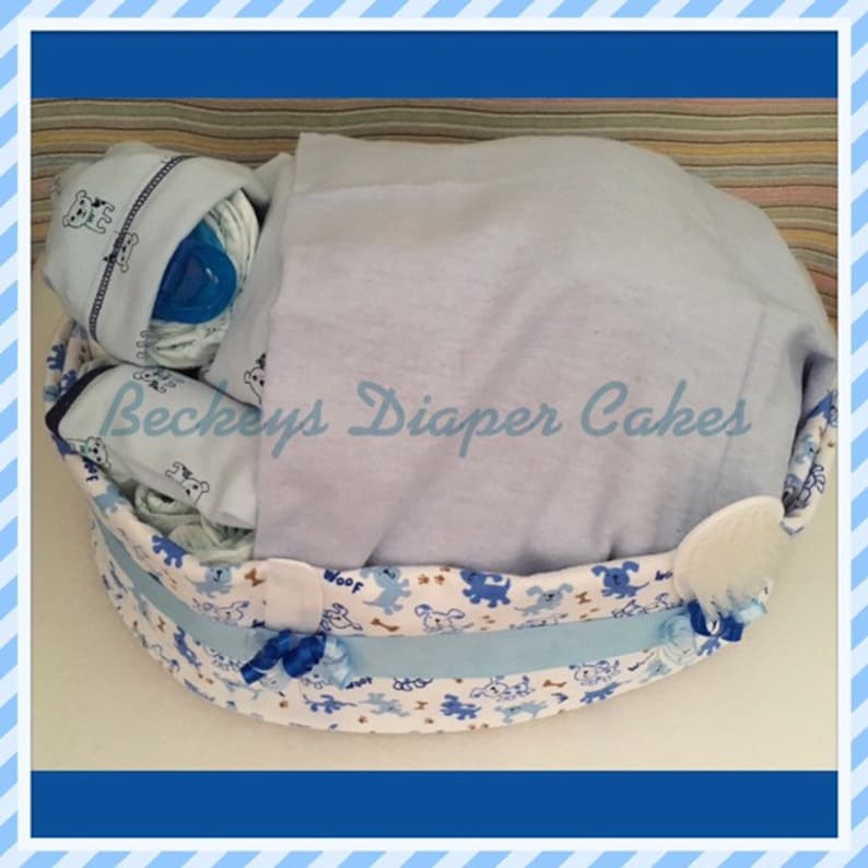 May include: A blue and white diaper cake decorated with a dog theme. The cake is in a basket with a blue blanket on top. The text "Beckeys Diaper Cakes" is visible on the blanket.