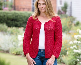 May include: A woman in a red, open-front cardigan with a scalloped edge, white top, and blue jeans. The cardigan is a bright red, possibly knitted or crocheted. The background shows a garden with greenery and a brick building, typical of a British garden.