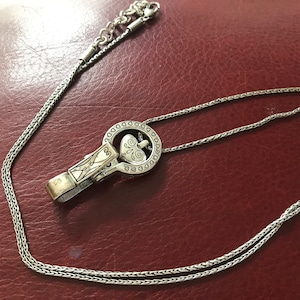 May include: A silver chain necklace with a heart-shaped pendant. The pendant is attached to a silver clip that is shaped like a circle with a heart in the center. The necklace is on a red leather surface.