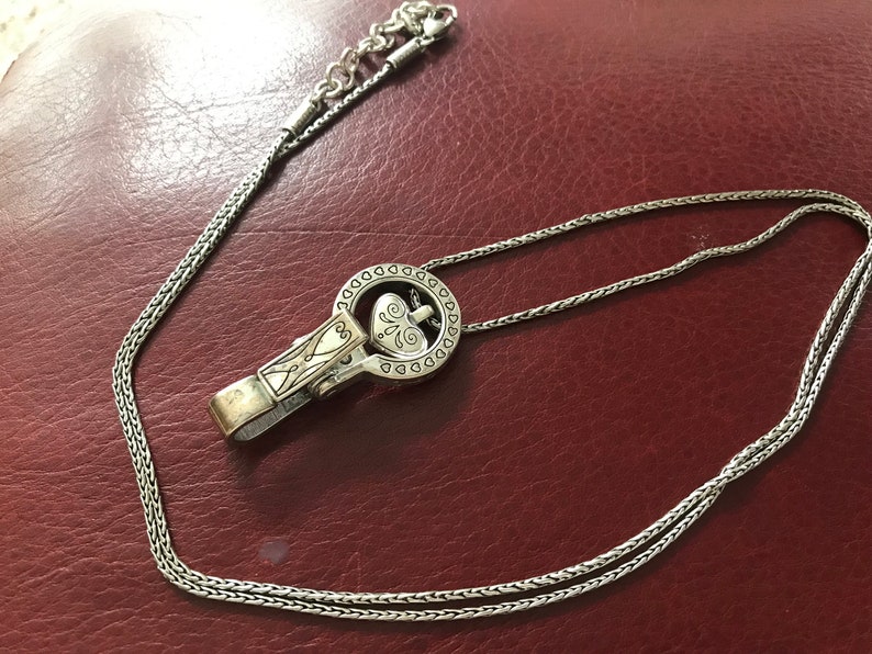 May include: A silver chain necklace with a heart-shaped pendant. The pendant is attached to a silver clip that is shaped like a circle with a heart in the center. The necklace is on a red leather surface.