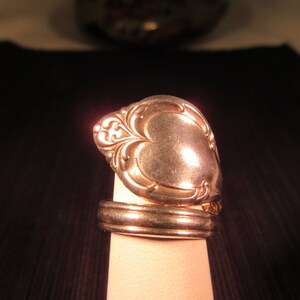 May include: A handcrafted gold-toned spoon ring. The ring features a decorative floral design on the spoon bowl and a simple band. The ring is displayed on a neutral-colored stand against a dark background.