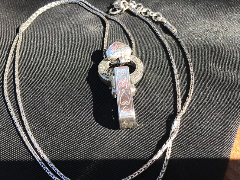 May include: A silver chain necklace with a heart-shaped pendant and a decorative clasp. The pendant and clasp feature intricate engraved designs.