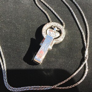 May include: A silver chain necklace with a heart-shaped pendant. The pendant has a detailed design and is attached to a clasp that looks like a key.