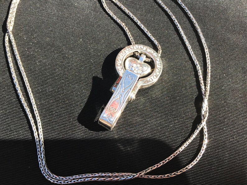 May include: A silver chain necklace with a heart-shaped pendant. The pendant has a detailed design and is attached to a clasp that looks like a key.