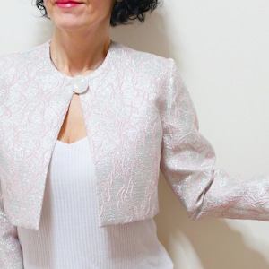 Puede incluir: Chaqueta bolero rosa claro con un patrón texturizado similar a flores. La chaqueta tiene un cuello redondo y un cierre de un solo botón blanco grande. Las mangas son largas y la chaqueta se usa sobre una camiseta blanca.