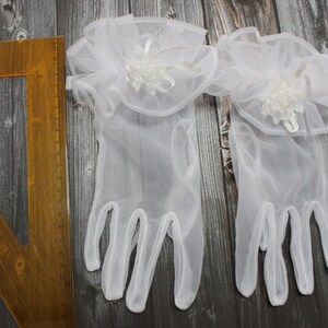 May include: A pair of white tulle gloves with a white lace ruffle and a white flower embellishment on each glove.