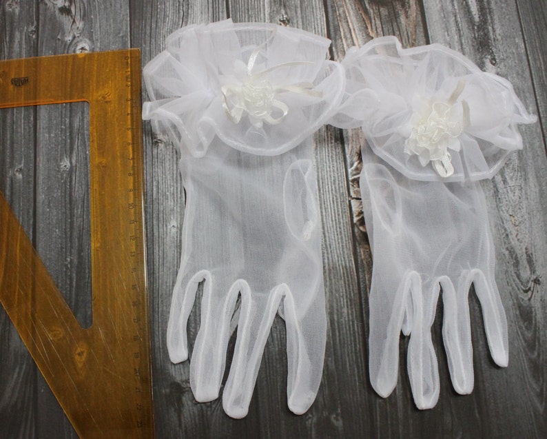 May include: A pair of white tulle gloves with a white lace ruffle and a white flower embellishment on each glove.