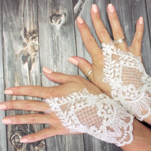 Könnte beinhalten: Weiße Spitzen-Fingerhandschuhe mit floralen und geometrischen Mustern. Die Handschuhe sind aus zarter Spitze gefertigt und weisen aufwendige Designs auf. Der Hintergrund ist eine Holzoberfläche.