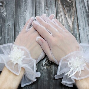 May include: A pair of white lace gloves with a ruffled trim and a white flower embellishment on each wrist.