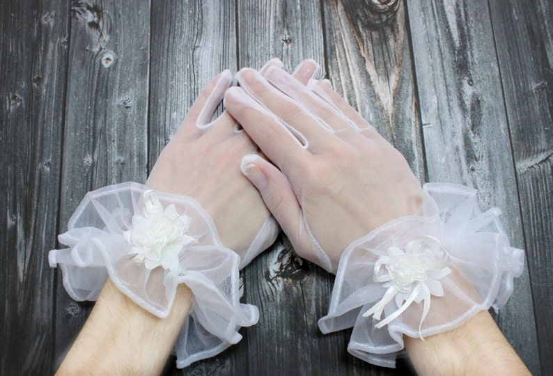 May include: A pair of white lace gloves with a ruffled trim and a white flower embellishment on each wrist.