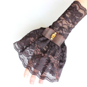 May include: A brown lace wrist cuff with a decorative gold button. The cuff is ruffled and has a scalloped edge.