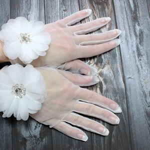 May include: A pair of white sheer gloves with a white flower embellishment on each wrist. The flowers have a rhinestone center.