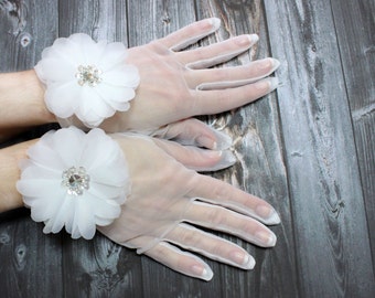 May include: A pair of white sheer gloves with a white flower embellishment on each wrist. The flowers have a rhinestone center.