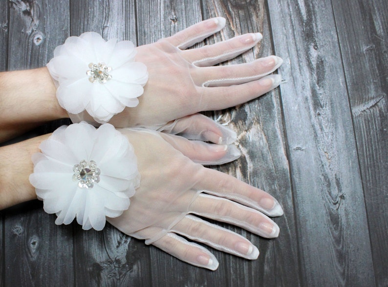 May include: A pair of white sheer gloves with a white flower embellishment on each wrist. The flowers have a rhinestone center.