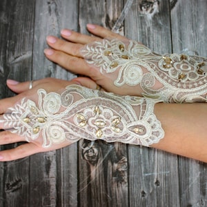 May include: A pair of ivory lace fingerless gloves with gold embellishments. The gloves are adorned with a delicate floral pattern and have a sheer, lightweight fabric.