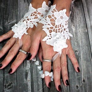 May include: A pair of white lace fingerless gloves with a white ribbon detail. The gloves are worn on a pair of hands with dark red nail polish. A white pearl necklace is visible on the right hand.