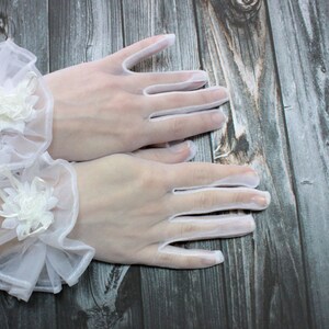 May include: A pair of white lace gloves with delicate floral embellishments. The gloves are sheer and have a wrist-length cuff.