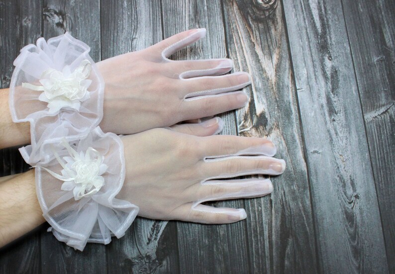 May include: A pair of white lace gloves with delicate floral embellishments. The gloves are sheer and have a wrist-length cuff.