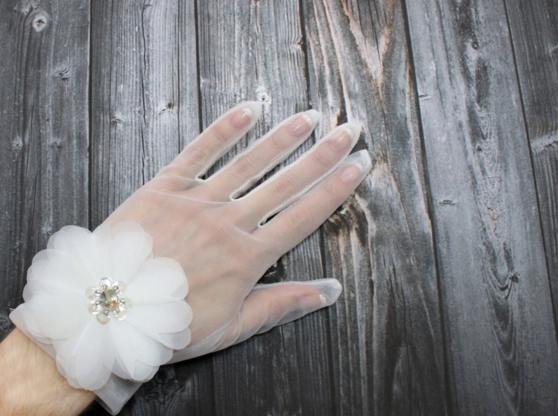 May include: White sheer gloves with a white flower embellishment on the wrist. The flower has a rhinestone center.