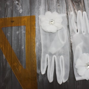 May include: A pair of white sheer gloves with a white flower embellishment on each glove. The flowers have a rhinestone center.