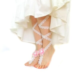 Puede incluir: Sandalias blancas de encaje para pies descalzos con detalles de flores rosas. Las sandalias se atan a las piernas con un patr&oacute;n de cruz.
