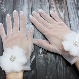 May include: A pair of white sheer gloves with a white flower embellishment on each wrist. The flowers have a rhinestone center.