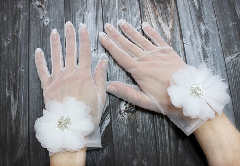 May include: A pair of white sheer gloves with a white flower embellishment on each wrist. The flowers have a rhinestone center.